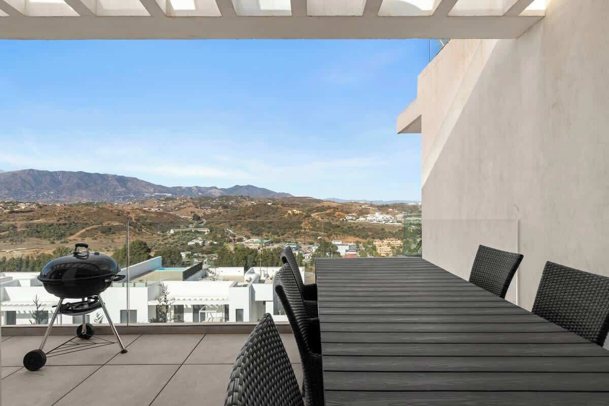 A rooftop patio with a grill, dining table, and chairs, overlooking a mountainous landscape.