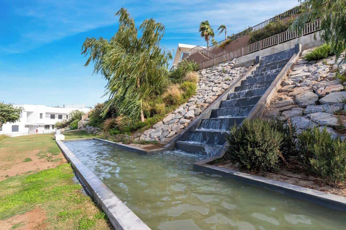 A water feature with cascading steps and a stone wall, surrounded by greenery and a clear sky.