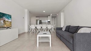 A modern living room with a grey sofa, a white coffee table, and a flat-screen TV on a white cabinet. There is a dining area with a white table and chairs, and a kitchen in the background. - Casa Banderas Pool & Gym by Costarentals - Ref 237 (La Cala de Mijas)