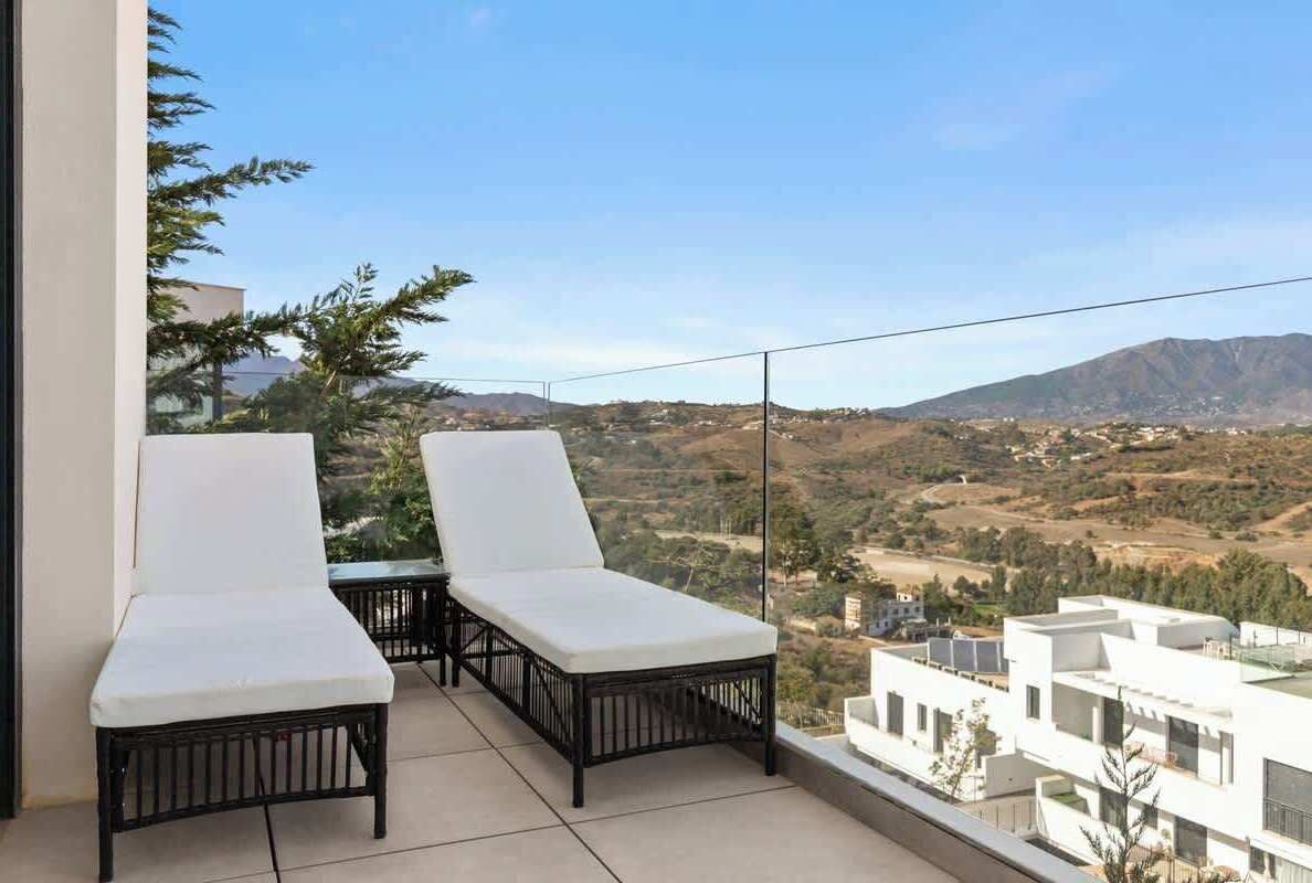 Ein Balkon mit zwei weißen, gepolsterten Stühlen und einem Tisch mit Blick auf eine hügelige Landschaft und Gebäude.