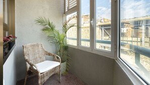 Een balkon met een rieten stoel, een potplant en uitzicht op gebouwen door het raam.