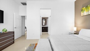 Une chambre moderne avec un grand lit, une table de chevet avec une lampe, un téléviseur à écran plat fixé au mur et un couloir menant à la salle de bain.