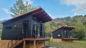 Deux cabanes dotées de terrasses en bois et d’une façade noire, situées dans une zone boisée sous un ciel dégagé.