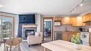 A modern living room with a fireplace, a dining area, and a kitchen with wooden cabinets and a microwave. - Lux 2BR/5BD Suite/Mountain View! (Canmore)