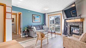 A modern living room with a sofa, a chair, a TV, and a fireplace. - Lux 2BR/5BD Suite/Mountain View! (Canmore)