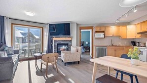 A modern living room with a fireplace, a dining area, and a kitchen. - Lux 2BR/5BD Suite/Mountain View! (Canmore)