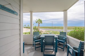 Un espace extérieur abrité avec une table et des chaises, une enseigne sur laquelle on peut lire « VIVEZ, AIMEZ, SURFEZ », et une vue sur la plage et l’océan.