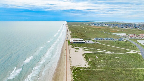 An aerial view of a coastal area with a beach, a small building complex, and a residential area with houses.