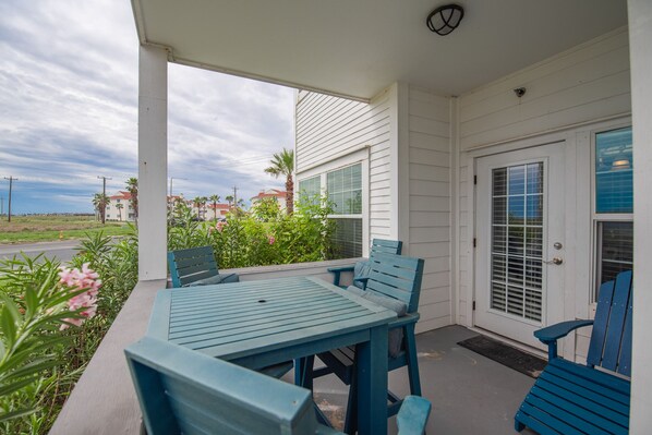 A covered patio with a table and chairs, a door with a window, and a view of a street with buildings and palm trees.