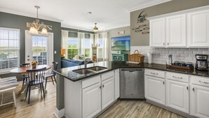 A kitchen with white cabinets, a black countertop, and a dishwasher. There is a dining area with a table and chairs, and a view of the outdoors through large windows.
