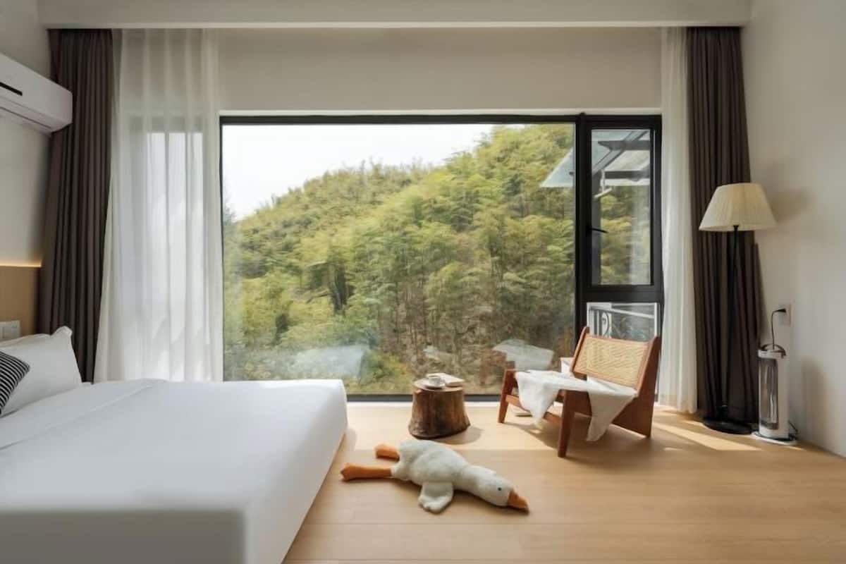 A hotel room with a bed, a chair, a lamp, and a view of a forest.