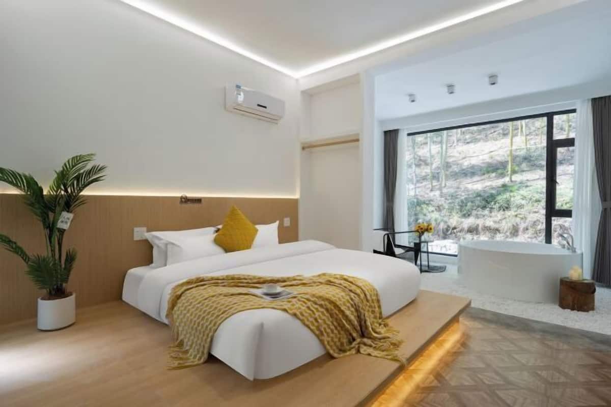 A modern hotel room with a bed, a small table with a vase of flowers, a potted plant, and a view of the outdoors.