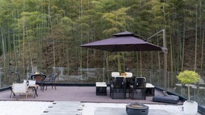Un espace extérieur aménagé avec une table et des chaises, entouré d’une rambarde en verre, offrant une vue sur une forêt de bambous.