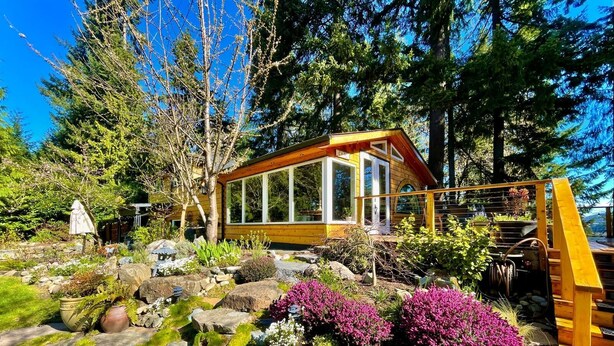 A wooden cabin with large windows, surrounded by a garden with rocks, moss, and flowering plants.