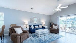 A spacious living room with a blue and white color scheme, featuring a blue sofa, two brown armchairs, a white coffee table, and a large window offering a view of the outside. - Premier Golf Location - Barefoot Resort! Family/dog friendly - 2 pools/beach! (North Myrtle Beach)