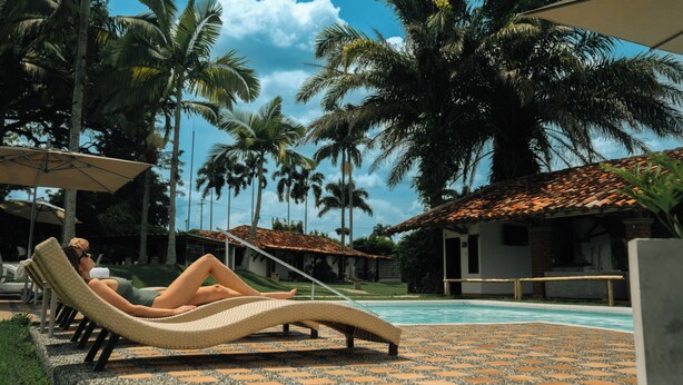 A poolside area with lounge chairs, palm trees, and a building with a tiled roof. - Hotel Rancho Eden (Armenia)