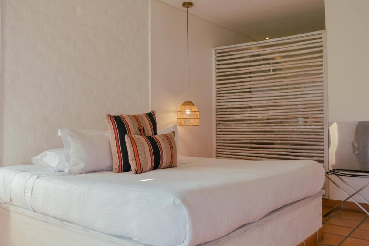 Quarto com cama, almofadas, luminária pendente e janela com persianas.