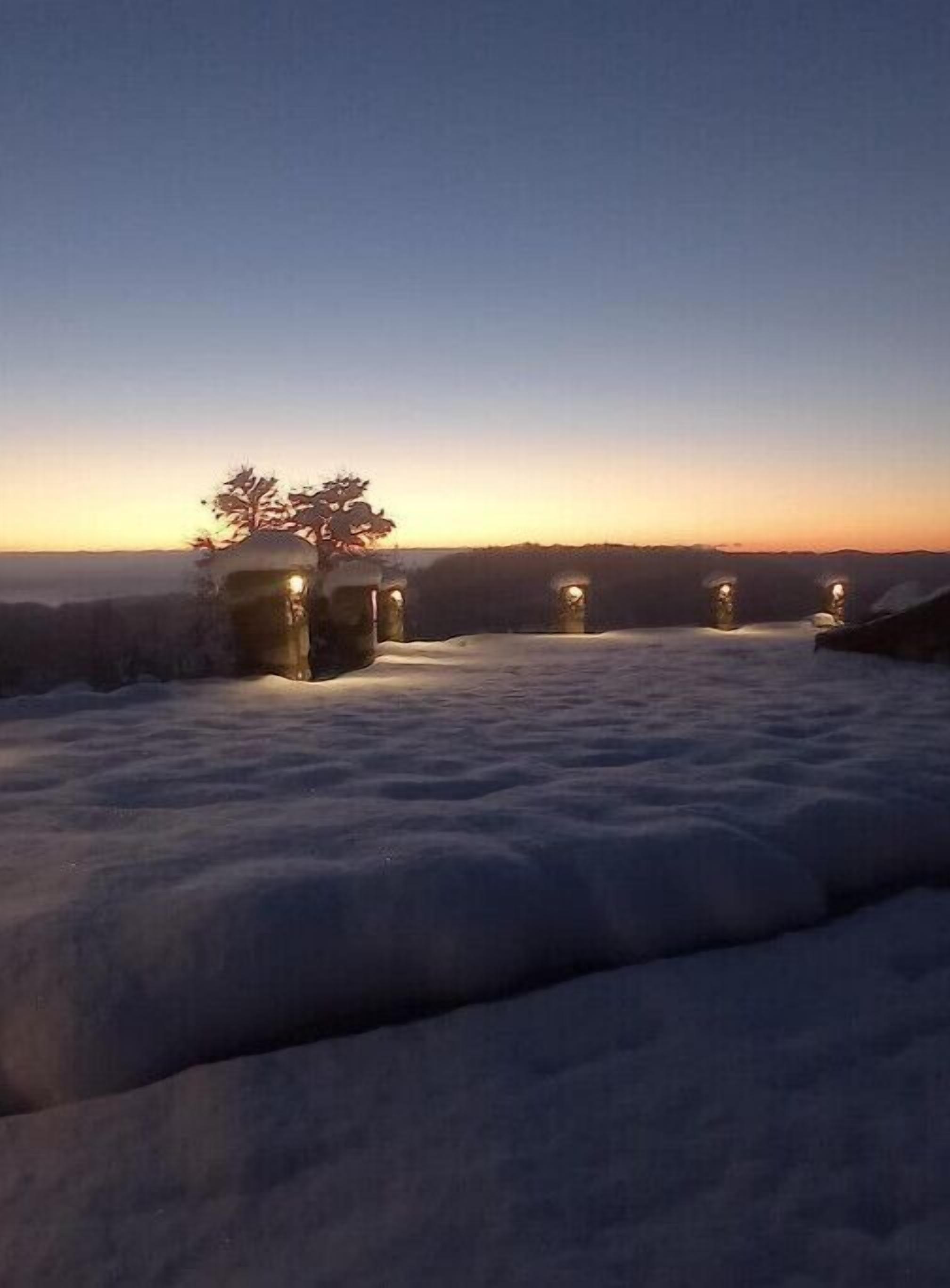 Ett snötäckt landskap i skymningen med en rad upplysta byggnader, troligen ett hotell eller ett semesterområde, och ett träd som avtecknas mot himlen.