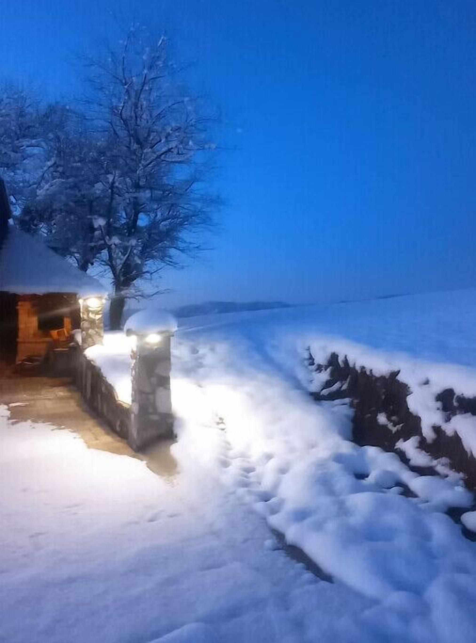 Un sentier enneigé mène à un mur de pierre et à un bâtiment dont les lumières sont allumées.