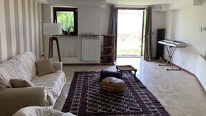 En stue med sofa, et mønstret tæppe, en radiator, et vindue med gardiner og et keyboardstativ.