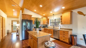 A spacious kitchen with wooden cabinets, a central island, stainless steel appliances, and a large window providing natural light. - Camp Manatee - Riverfront Cabin, Stunning Views, Pet-Friendly, Hot tub + Kayaks! (Fort White)