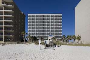 Un hotel frente a la playa con varios pisos, balcones y una zona de arena con palmeras.