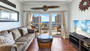 Een woonkamer met een leren bank, een rieten poef, een flatscreen televisie en een balkon met uitzicht op gebouwen en de zee.