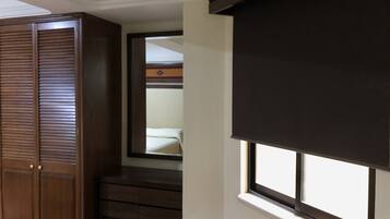 Una habitación con un armario de madera, una cama con cabecera estampada y un ventanal con persiana oscura.