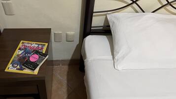 Una cama con sábanas blancas, una mesita de noche con una revista e interruptor de luz en la pared.