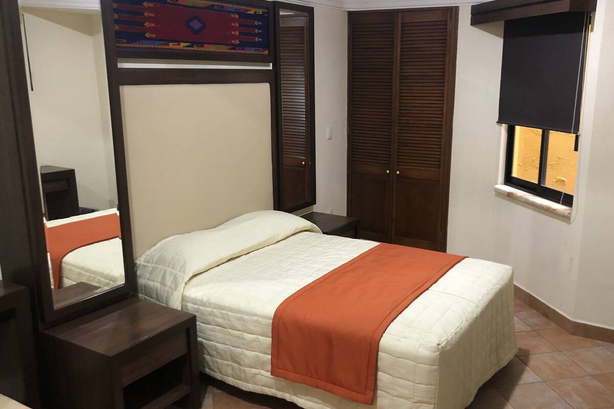Un dormitorio con cama, mesita de noche, espejo, armario y ventana con persiana.