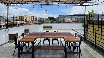 Una terraza en la azotea con una mesa y sillas de madera, una pérgola con luces colgantes, y vistas a un paisaje urbano y montañas al fondo.