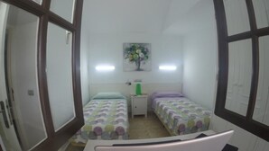 A room with two beds, a desk with a laptop, a nightstand, a wall-mounted painting, and a window with blinds.