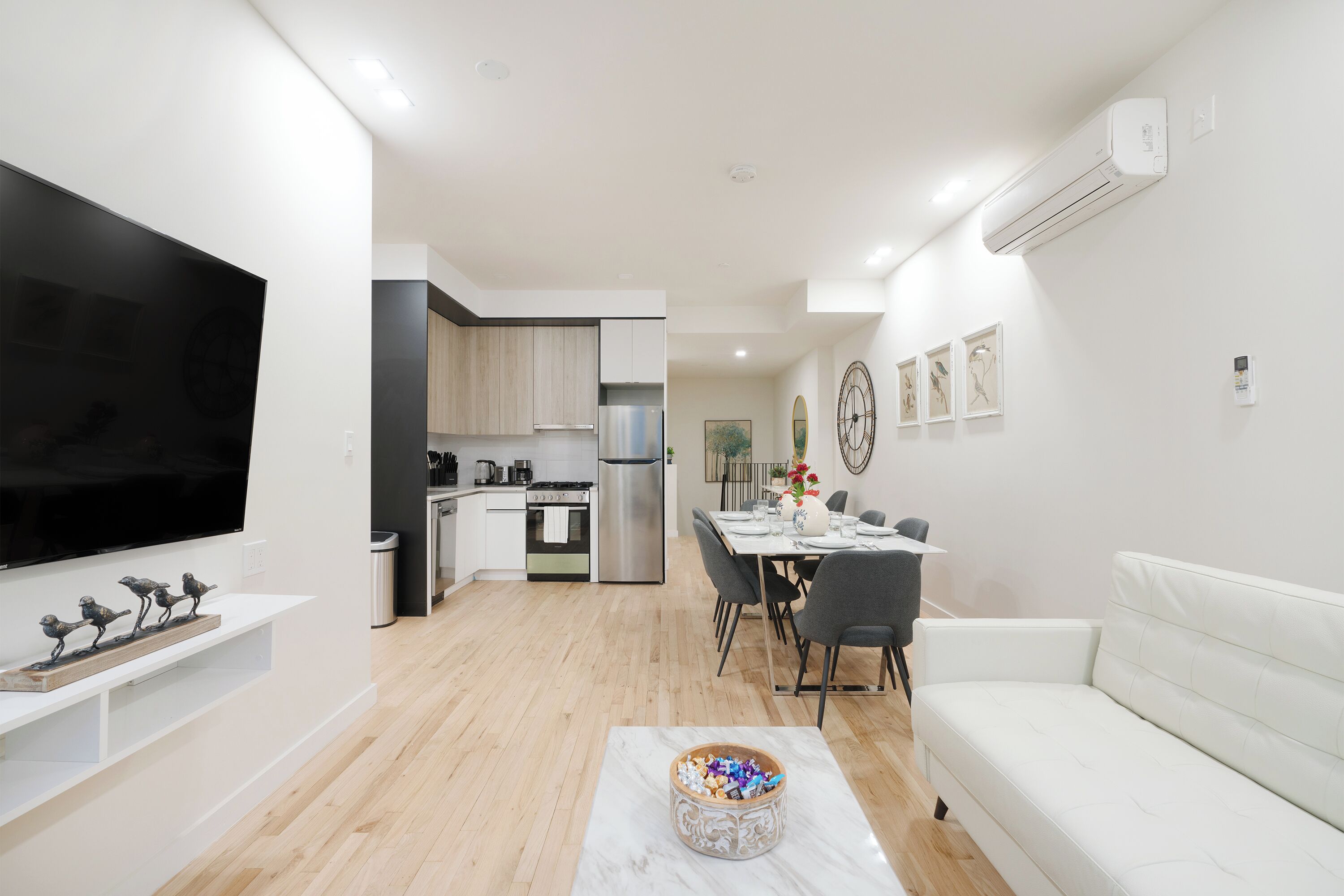 Uma sala de estar moderna com televisão de tela plana, um sofá branco, uma mesa de jantar com cadeiras e uma cozinha ao fundo.