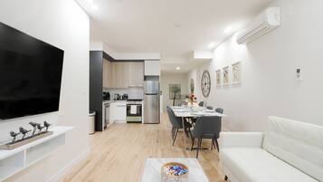 Uma sala de estar moderna com televisão de tela plana, um sofá branco, uma mesa de jantar com cadeiras e uma cozinha ao fundo.