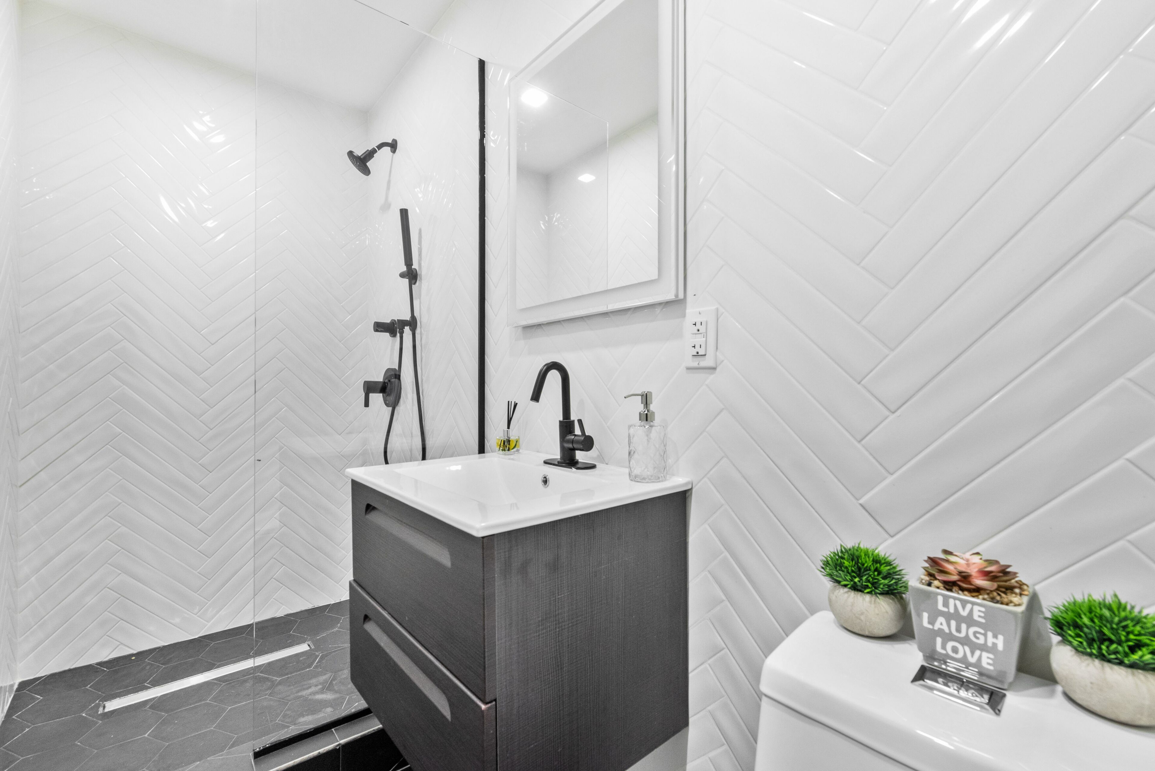 A modern bathroom with a white herringbone tile wall, a grey cabinet, a white sink, a black faucet, a handheld showerhead, a toilet, and two potted plants with a motivational sign.