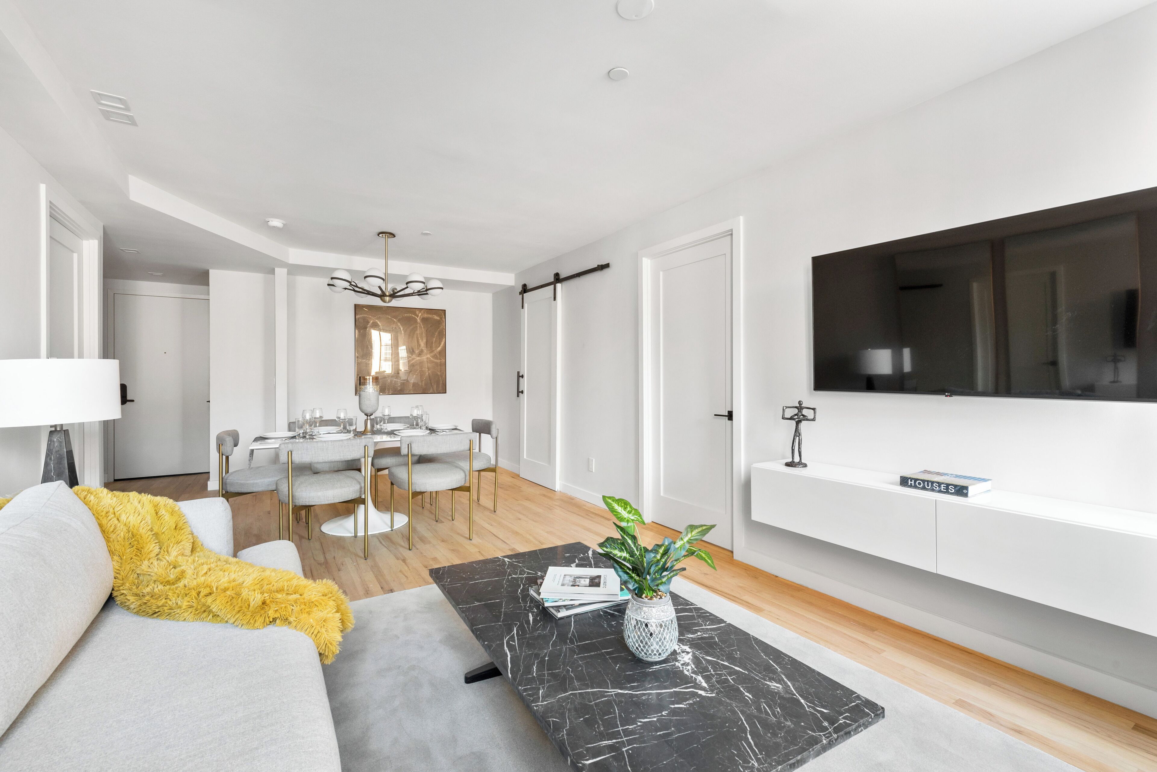 A modern living room with a sofa, a coffee table, a dining area with a table and chairs, and a TV mounted on the wall.