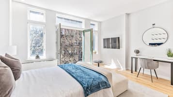 Un dormitorio moderno con una cama grande, un escritorio y una silla. Hay una ventana con vistas a árboles y una puerta que da a otra habitación.