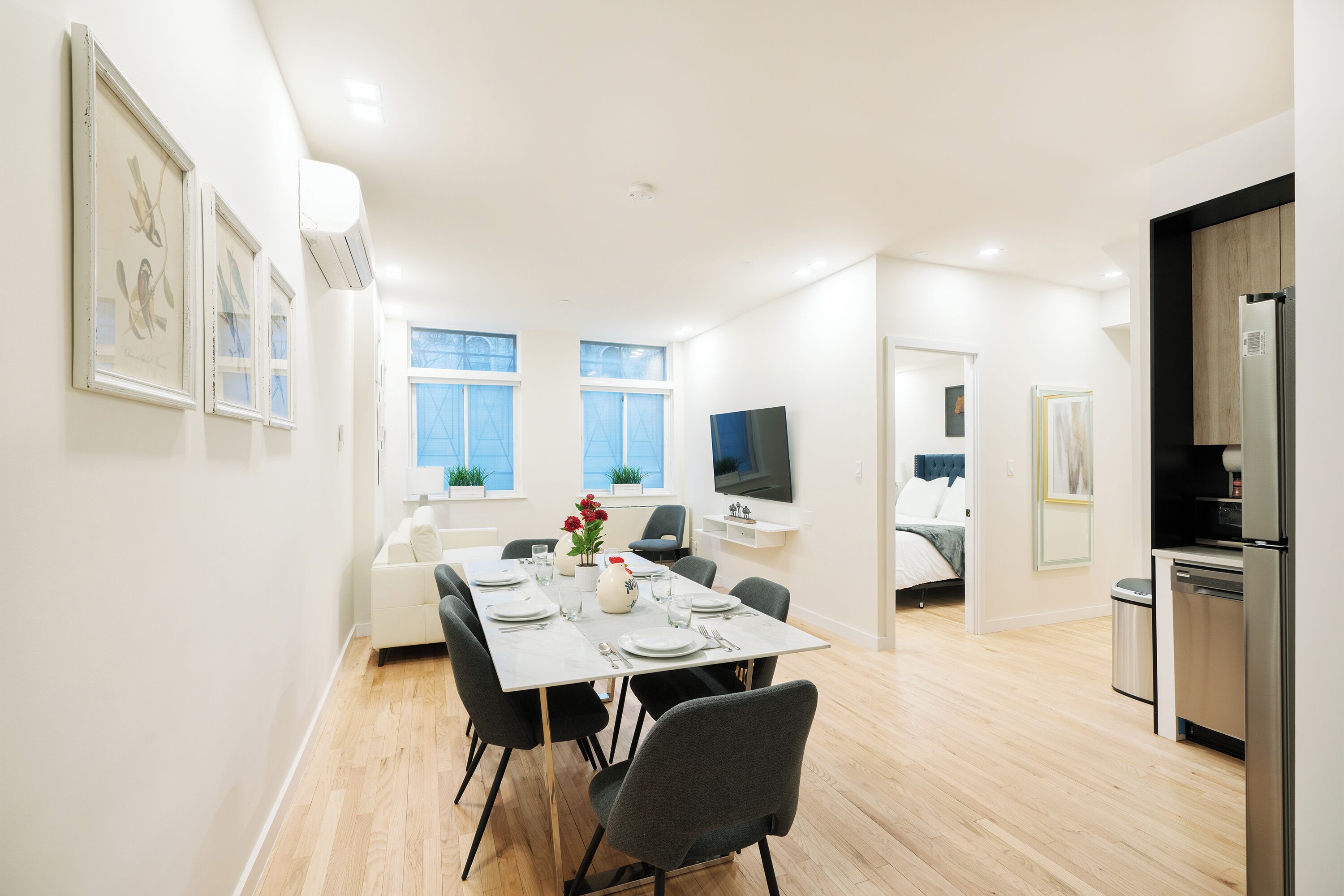 Un comedor moderno y luminoso con una mesa blanca dispuesta para cuatro personas, rodeada de sillas grises. Hay obras de arte enmarcadas en las paredes, un televisor de pantalla plana y una zona de cocina con refrigerador y fregadero.