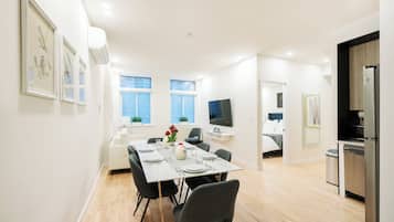 Un comedor moderno y luminoso con una mesa blanca dispuesta para cuatro personas, rodeada de sillas grises. Hay obras de arte enmarcadas en las paredes, un televisor de pantalla plana y una zona de cocina con refrigerador y fregadero.