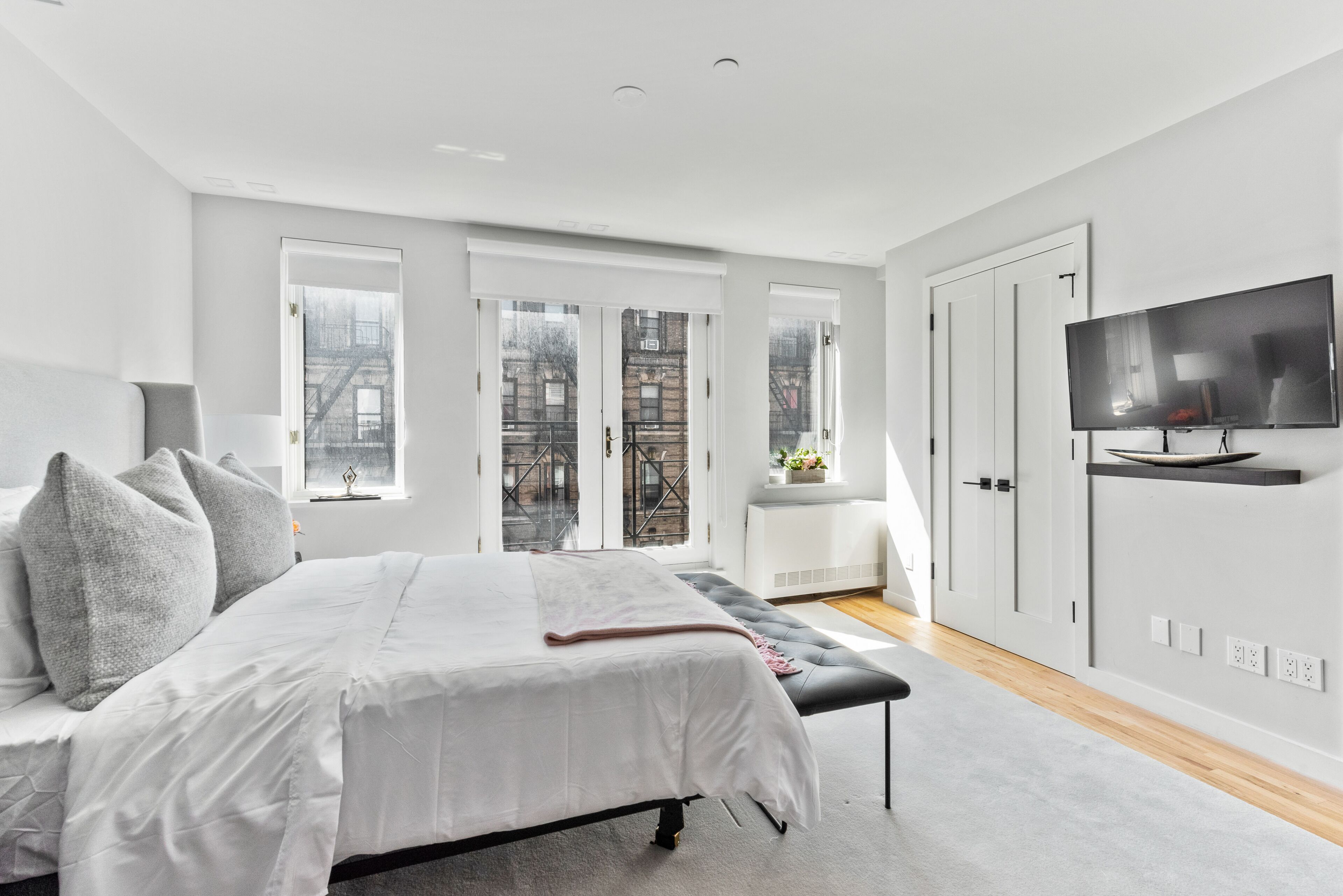 A modern bedroom with a large bed, a bench, a TV, and a view of the city through large windows.