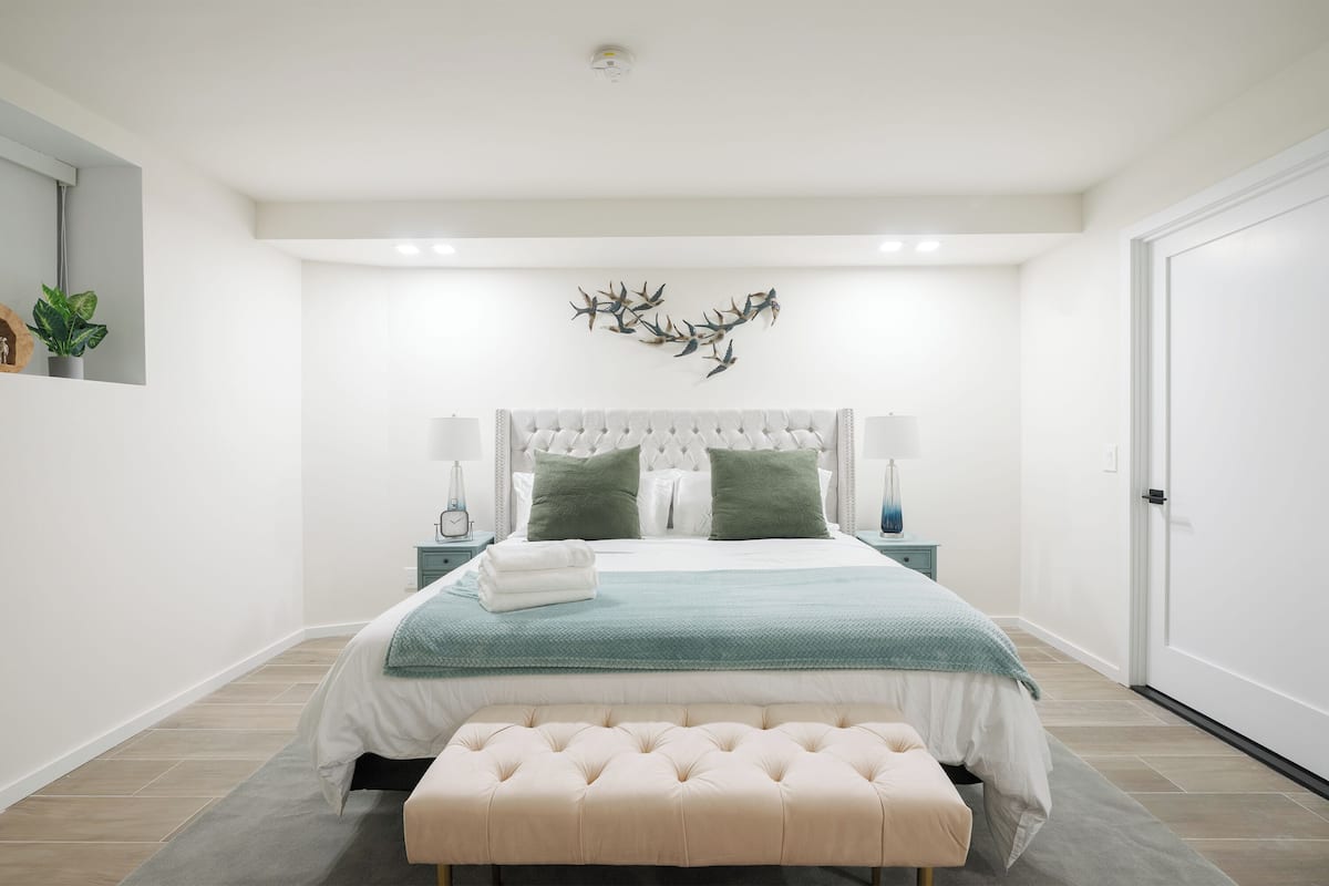 A modern bedroom with a large bed, a bench, a wall-mounted deer head sculpture, and a small plant on the left.