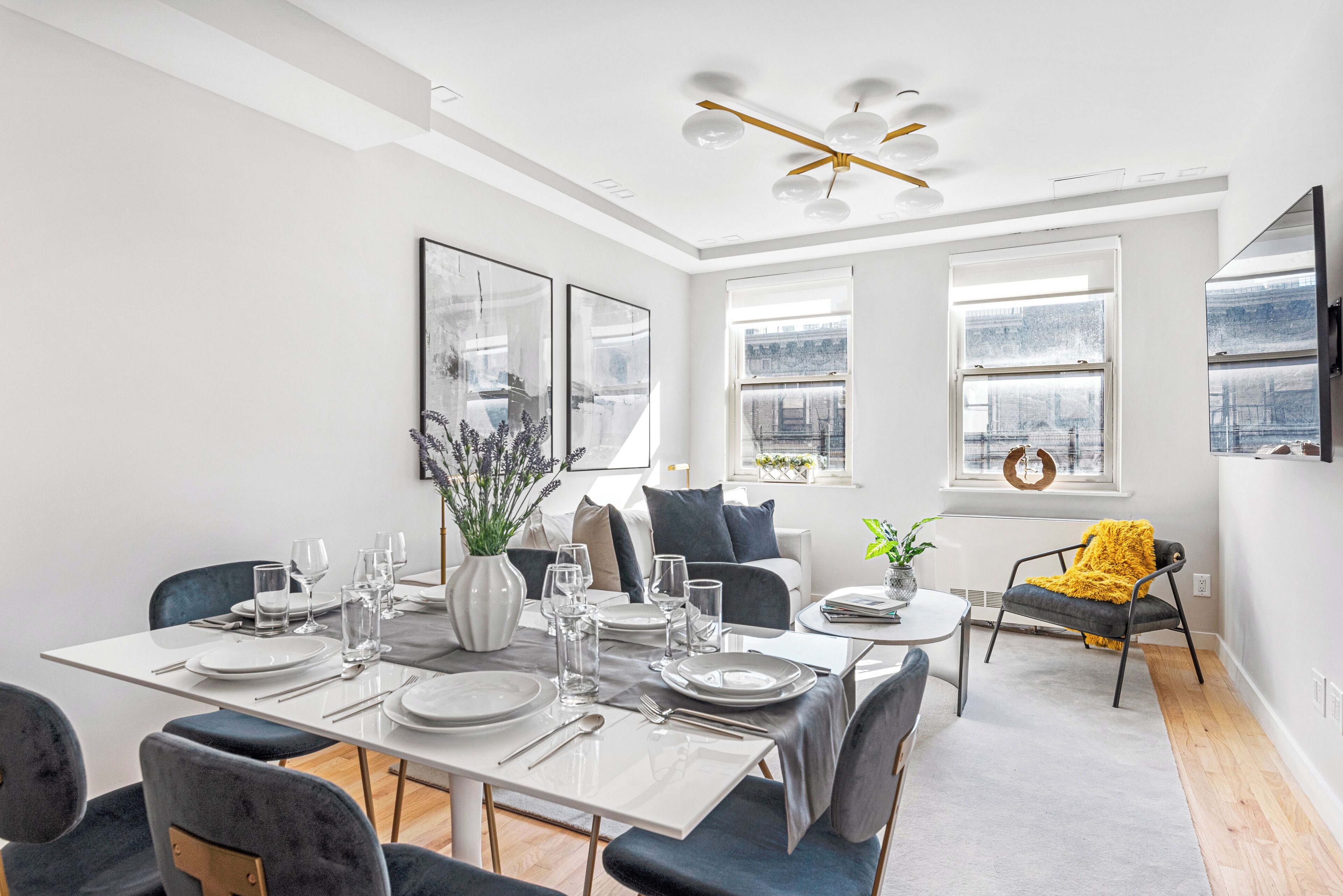 Un espace de salle à manger moderne avec une table blanche dressée pour six personnes, entourée de chaises foncées. On y trouve deux fenêtres à cadres blancs, un ventilateur de plafond et un plaid jaune sur une chaise.