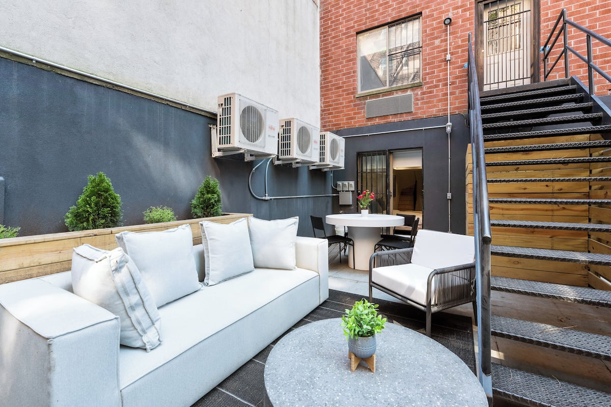 An outdoor seating area with a sofa, chairs, and a small table with a plant. There are air conditioning units mounted on the wall, a staircase, and a door leading to an interior space.