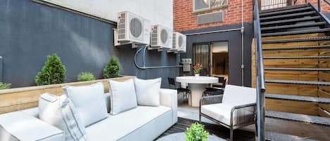 An outdoor seating area with a sofa, chairs, and a small table with a plant. There are air conditioning units mounted on the wall, a staircase, and a door leading to an interior space.
