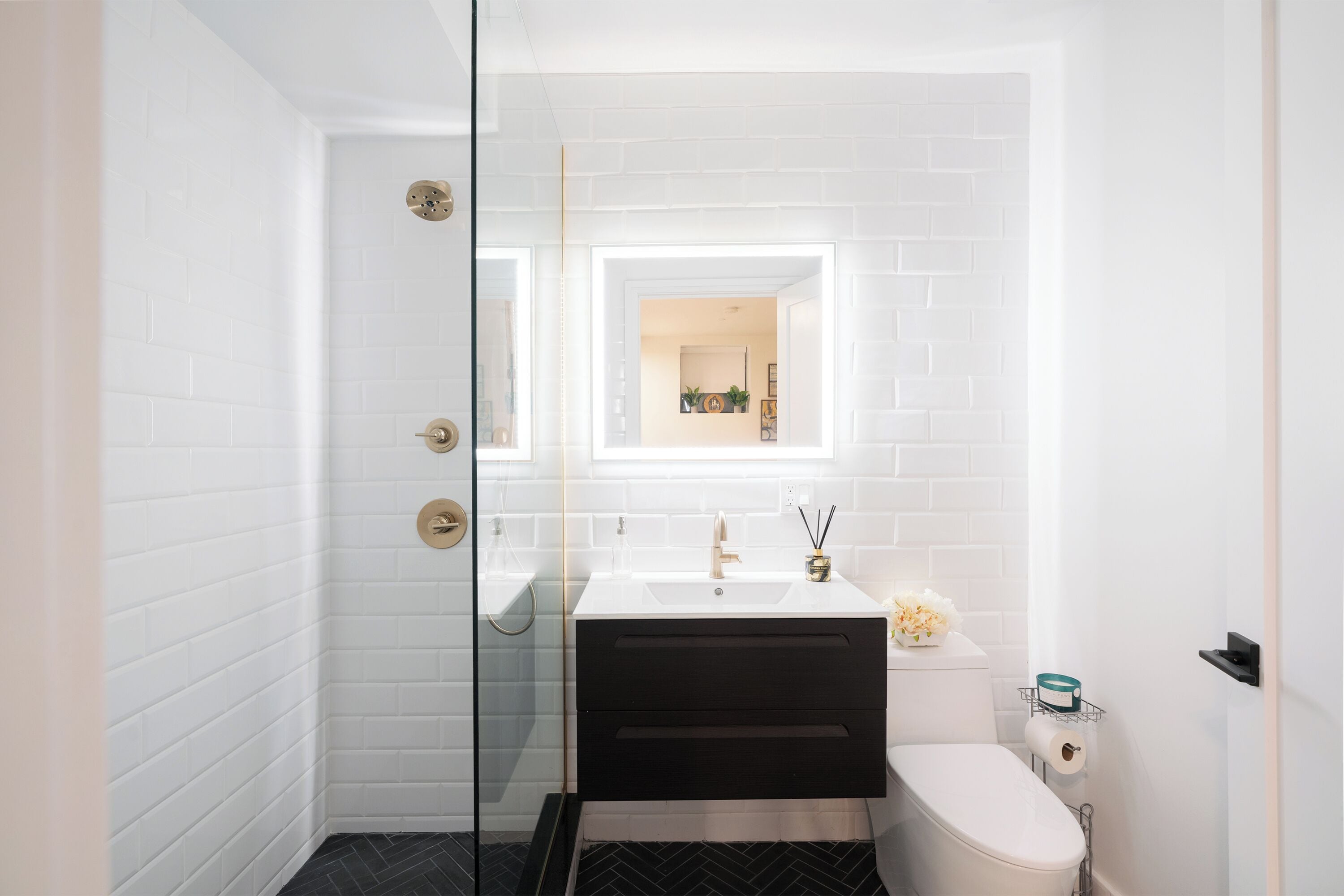 Un baño moderno con azulejos blancos tipo metro, un tocador blanco y negro, una cabina de ducha de vidrio y un inodoro blanco.
