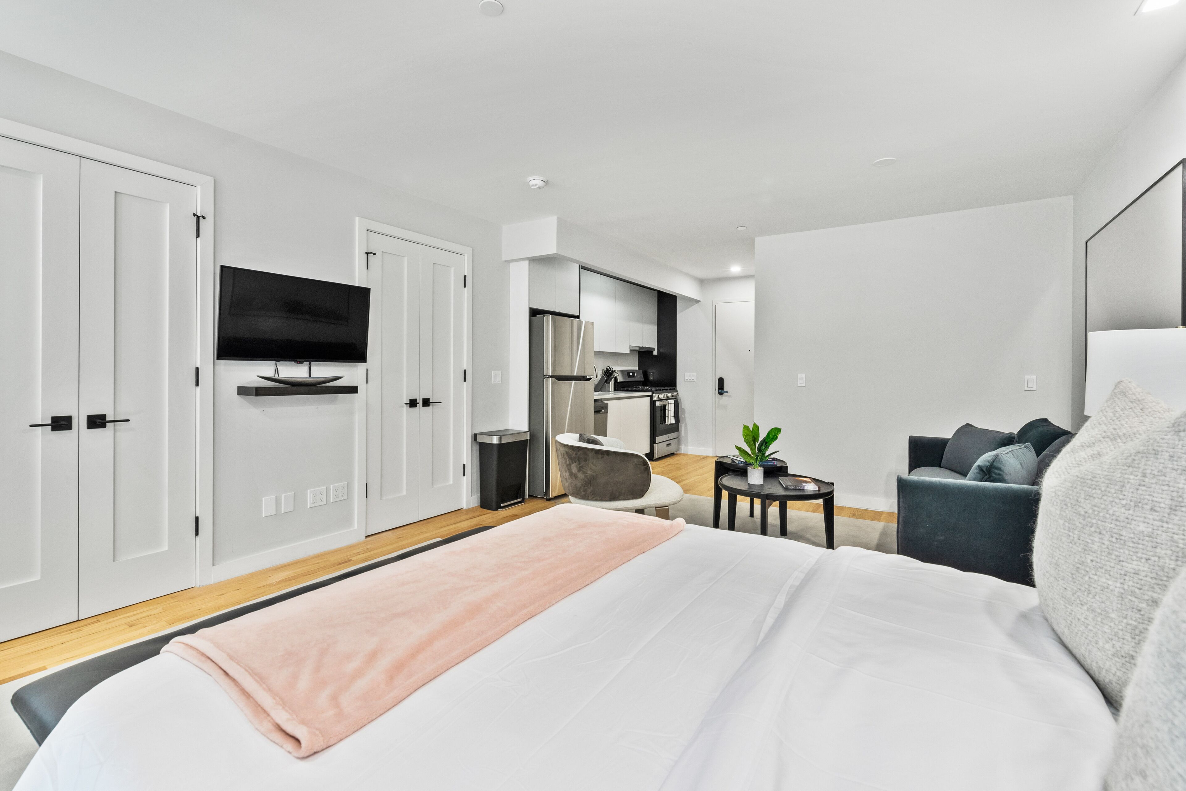 Habitació d'hotel moderna amb un llit gran, un televisor fixat a la paret, una cuineta i una zona d'estar amb una cadira i una taula petita.