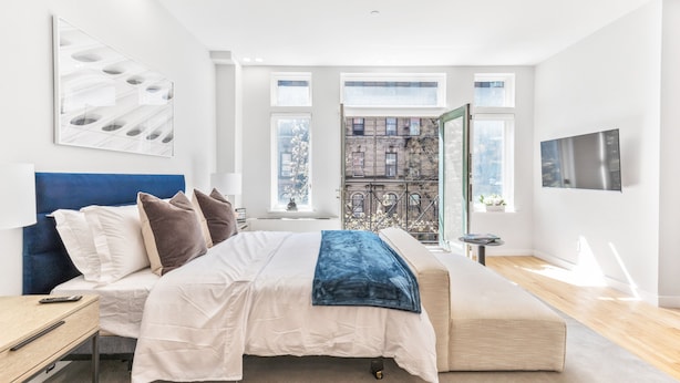 A modern bedroom with a large bed, a bedside table, a nightstand, a window with a city view, and a mounted TV. - The Sixty Two (New York)