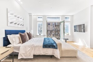 Un dormitorio moderno con una cama grande, una mesita de noche y un televisor montado. Se aprecia una ventana con vistas a la ciudad.
