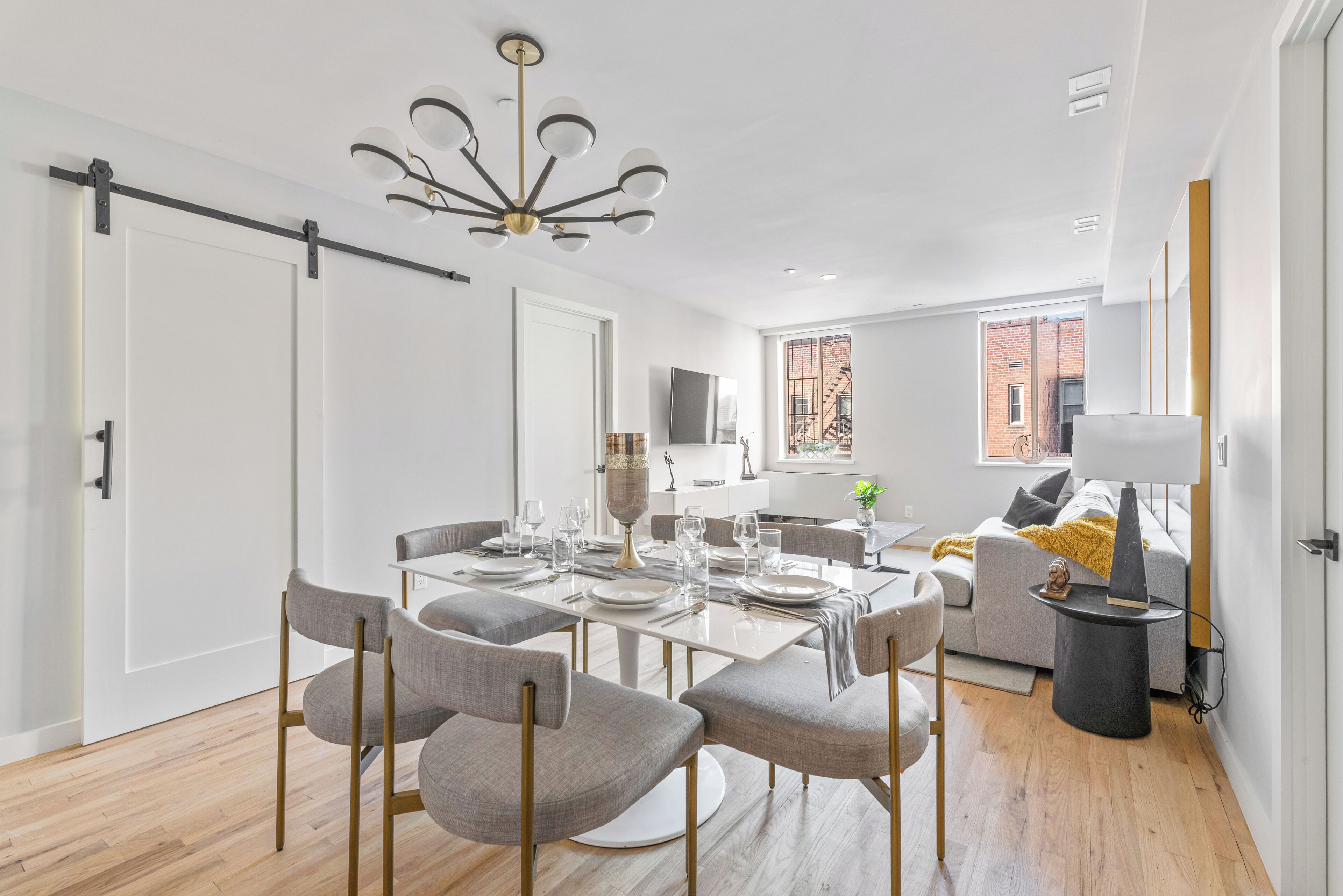 A modern dining area with a white table set for six, surrounded by grey upholstered chairs. A contemporary chandelier hangs from the ceiling, and a grey sofa is visible in the background.