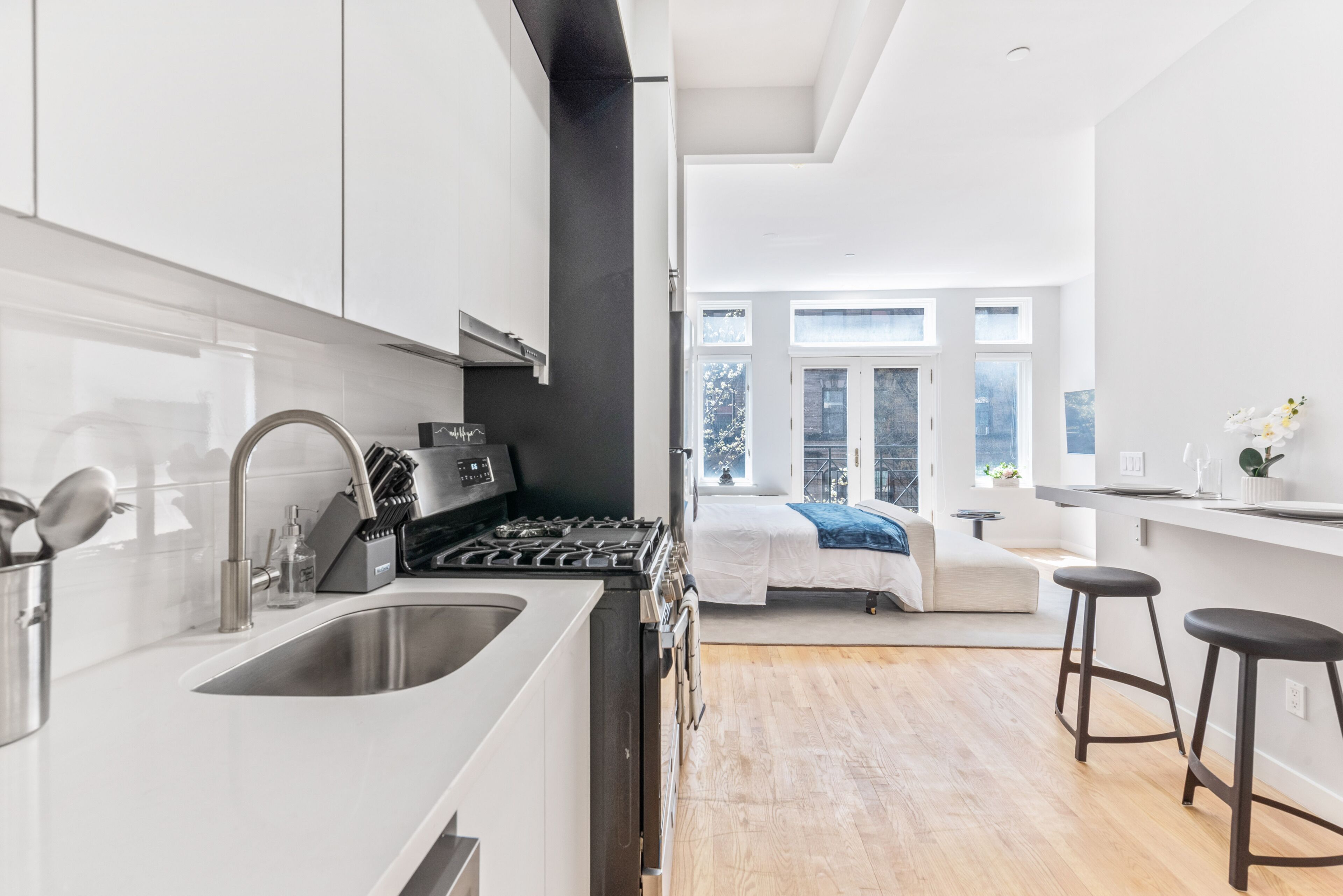 A modern kitchen with a large sink, stove, and black cabinets. There is a bed with a blue blanket and a window with a view of trees.